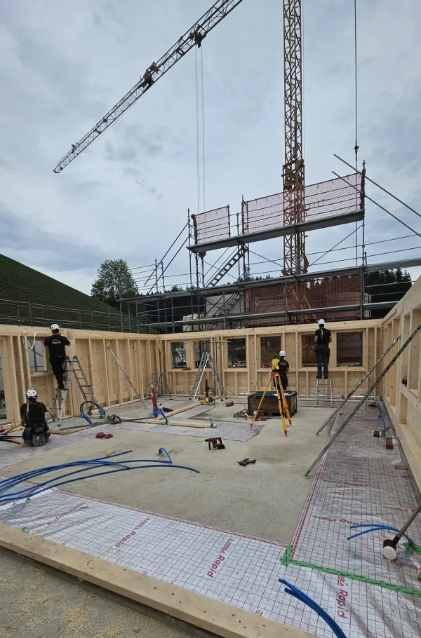 Holzbau im Rohbau mit Baukran an einem grünen Hang, mit bewaldeten Hügeln und schneebedeckten Bergen im Hintergrund.