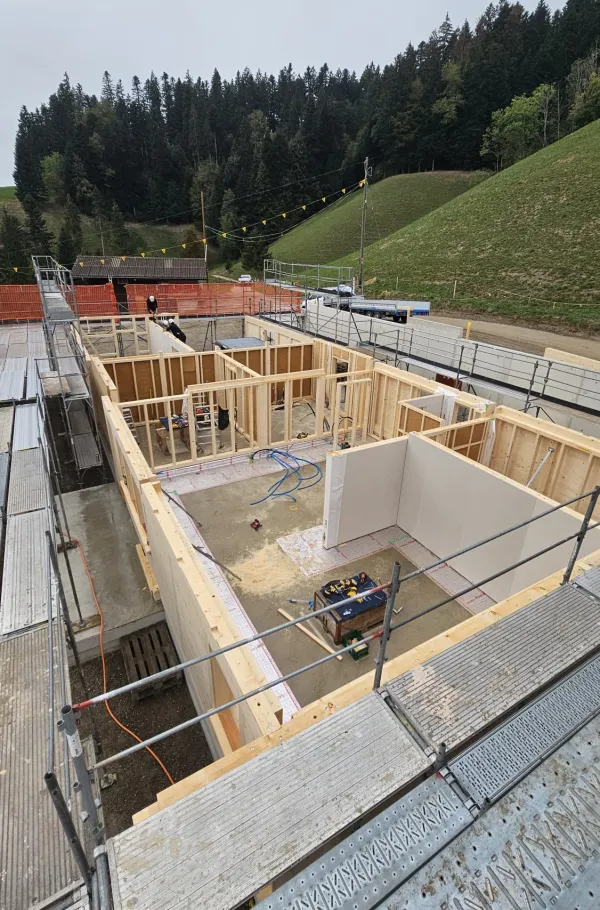 Holzbau im Rohbau mit Baukran an einem grünen Hang, mit bewaldeten Hügeln und schneebedeckten Bergen im Hintergrund.