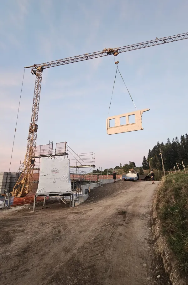 Holzbau im Rohbau mit Baukran an einem grünen Hang, mit bewaldeten Hügeln und schneebedeckten Bergen im Hintergrund.