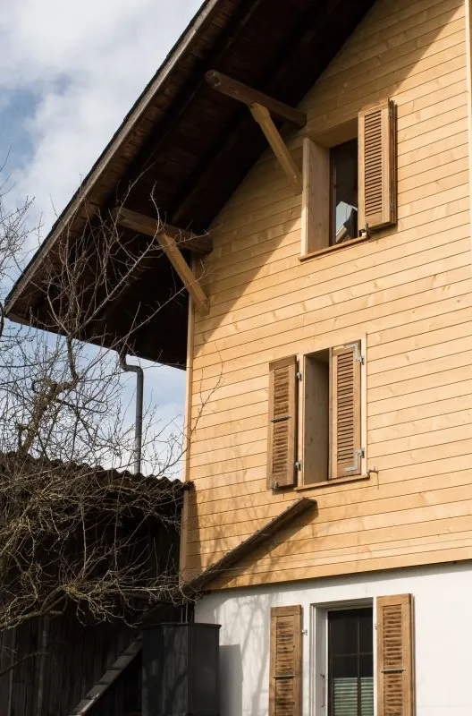 Referenzprojekt der Zimmerei Hirschi AG: Wohnhaus mit umfassend erneuerter Holzfassade, eingebettet in die typische Landschaft des Emmentals mit Wiesen und bewaldetem Hang.