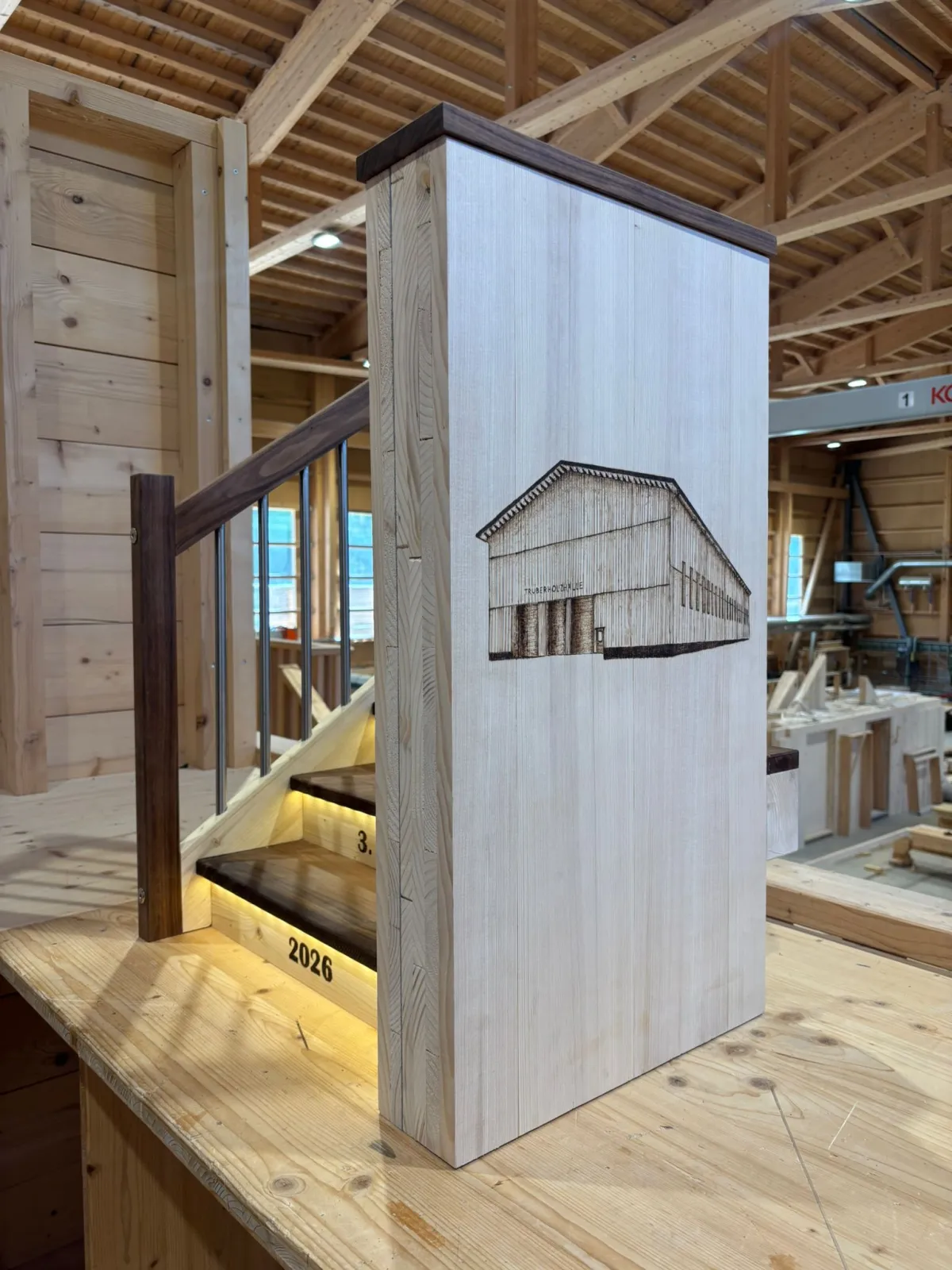Eine Lernende im Holzbau lehnt lächelnd an einem Holzgeländer und steht neben einem beleuchteten Treppenmodell mit der Aufschrift ‚Treppenmodell, 3. Lehrjahr, 2026‘. Die Szene befindet sich in einer grossen, modernen Zimmerei-Werkhalle mit sichtbarer Holzkonstruktion, Dachbalken und verschiedenen Holzbauelementen im Hintergrund.
