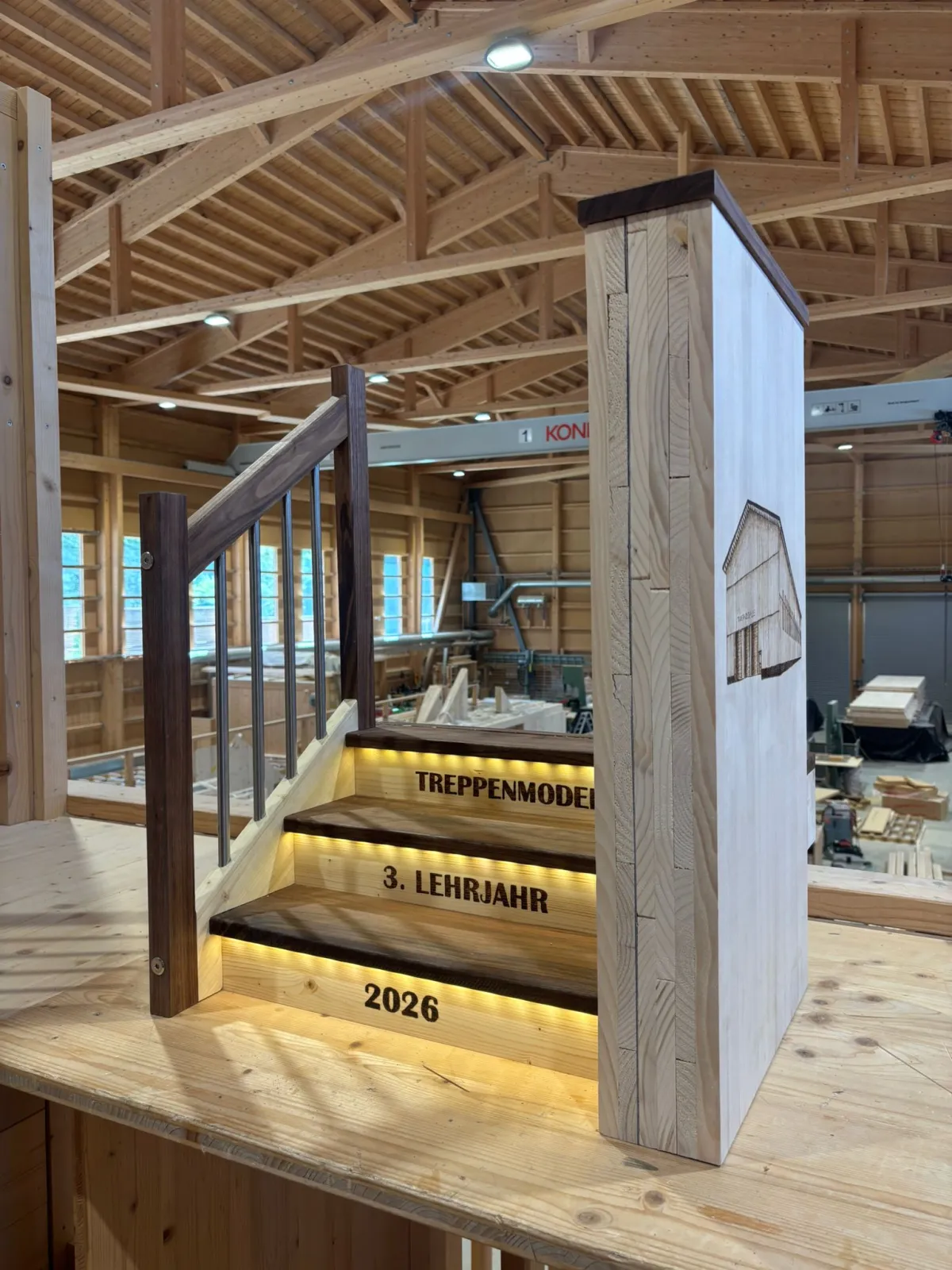 Eine Lernende im Holzbau lehnt lächelnd an einem Holzgeländer und steht neben einem beleuchteten Treppenmodell mit der Aufschrift ‚Treppenmodell, 3. Lehrjahr, 2026‘. Die Szene befindet sich in einer grossen, modernen Zimmerei-Werkhalle mit sichtbarer Holzkonstruktion, Dachbalken und verschiedenen Holzbauelementen im Hintergrund.