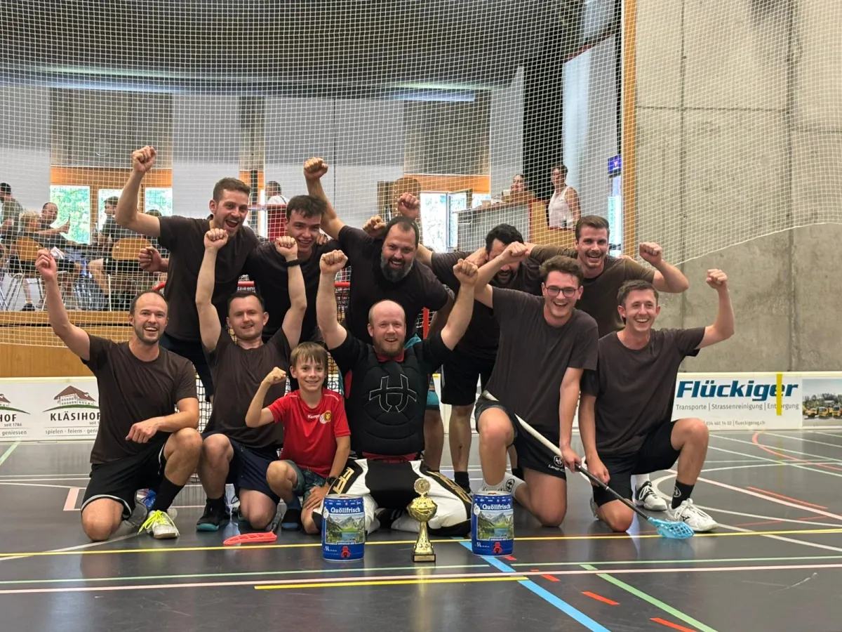 Eine Unihockey-Mannschaft feiert gemeinsam mit einem Kind und einem Torhüter auf dem Hallenboden einen Turniersieg. Vor der Gruppe stehen ein Pokal und zwei Getränkepreise.