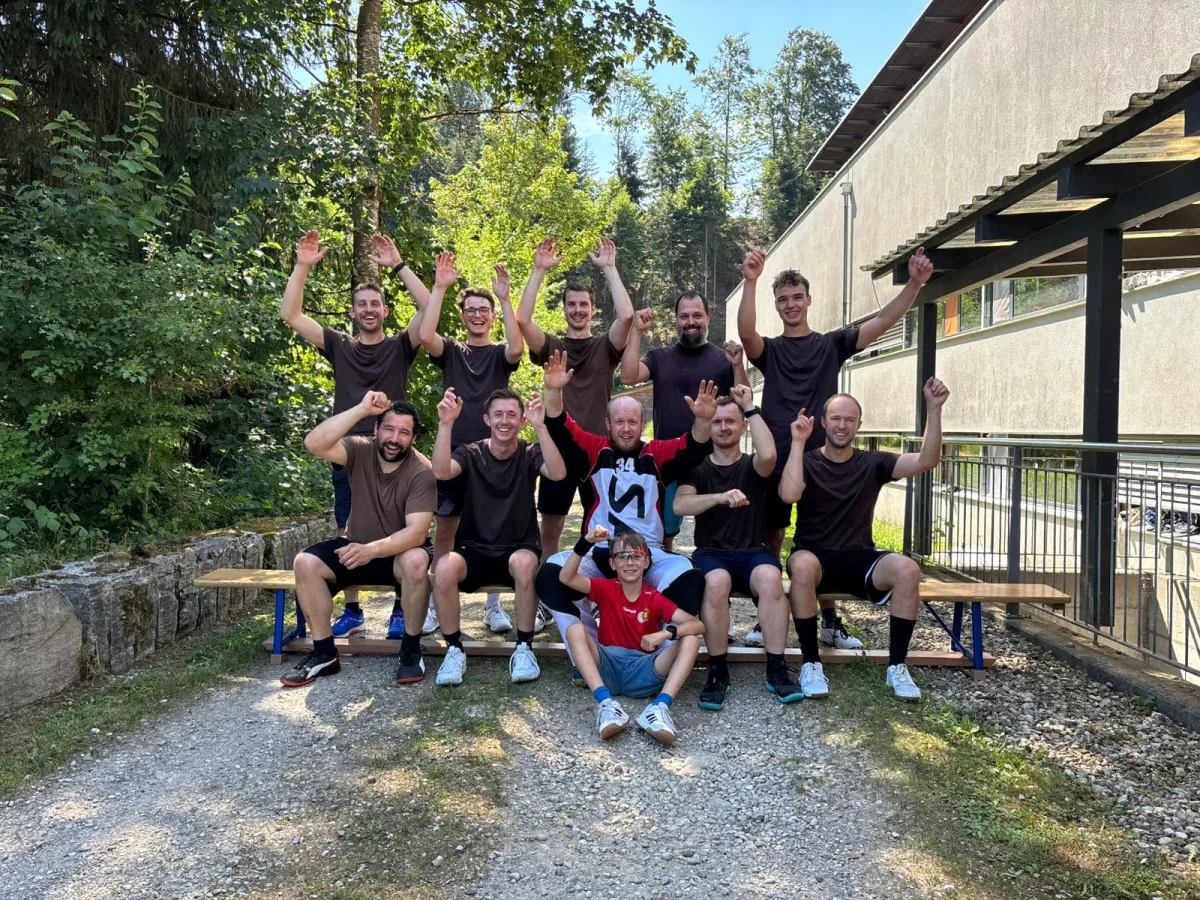 Eine Unihockey-Mannschaft feiert gemeinsam mit einem Kind und einem Torhüter auf dem Hallenboden einen Turniersieg. Vor der Gruppe stehen ein Pokal und zwei Getränkepreise.