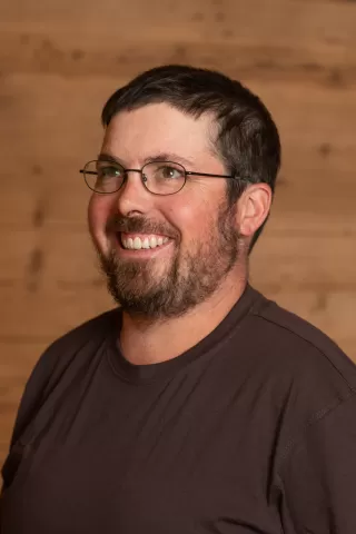 Mitarbeiterportrait / Foto mit braunem T-Shirt oder schwarzem Polo Shirt mit Zimmerei Hirschi Logo. Strahlend und lachend nach oben schauend oder direkt in die Kamera.