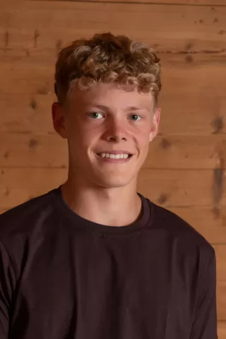 Porträtfoto eines jungen Mannes mit lockigem, blondem Haar und freundlichem Lächeln vor einem rustikalen Holz-Hintergrund. Er trägt ein dunkelbraunes T-Shirt und blickt direkt in die Kamera.