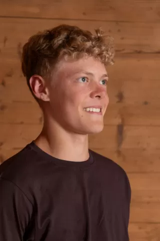 Profilfoto eines jungen Mannes mit lockigem, blondem Haar vor einer Holz-Wand. Er trägt ein dunkelbraunes T-Shirt und blickt mit einem freundlichen Ausdruck leicht nach rechts.