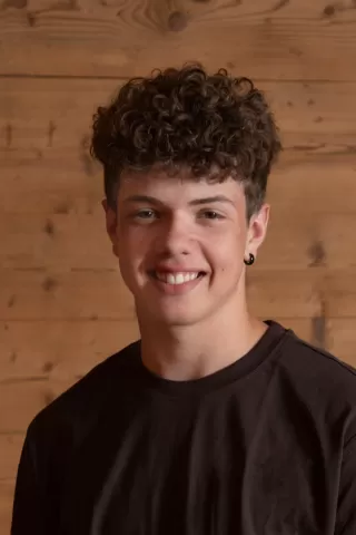 Porträtfoto eines jungen Mannes mit lockigem, braunem Haar und Ohrring. Er trägt ein dunkelbraunes T-Shirt und lächelt freundlich in die Kamera. Der Hintergrund besteht aus einer rustikalen Holz-Wand.