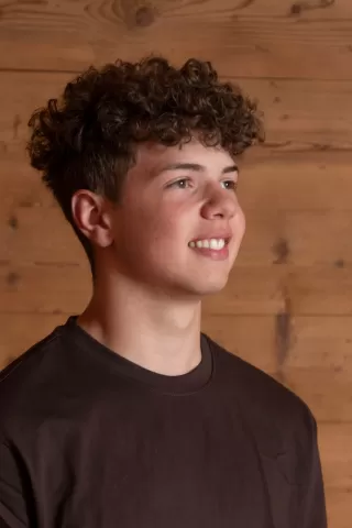 Profilfoto eines jungen Mannes mit lockigem Haar, der leicht nach rechts schaut und freundlich lächelt. Er trägt ein dunkelbraunes T-Shirt und steht vor einer Wand aus Naturholz.
