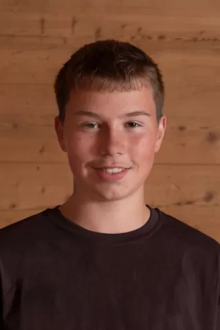 Porträtfoto eines jungen Mannes mit kurzem, braunem Haar und einem dezenten Lächeln. Er trägt ein dunkelbraunes T-Shirt und steht vor einer hölzernen Hintergrundwand.