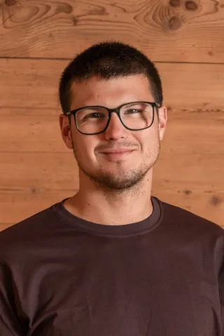 Porträt eines Mannes mit kurzen dunklen Haaren und Brille. Er trägt ein dunkles T-Shirt und blickt lächelnd in die Kamera; im Hintergrund ist eine helle Holzverkleidung zu sehen.