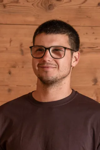 Porträt eines Mannes mit kurzen dunklen Haaren und Brille. Er trägt ein dunkles T-Shirt und blickt lächelnd in die Kamera; im Hintergrund ist eine helle Holzverkleidung zu sehen.