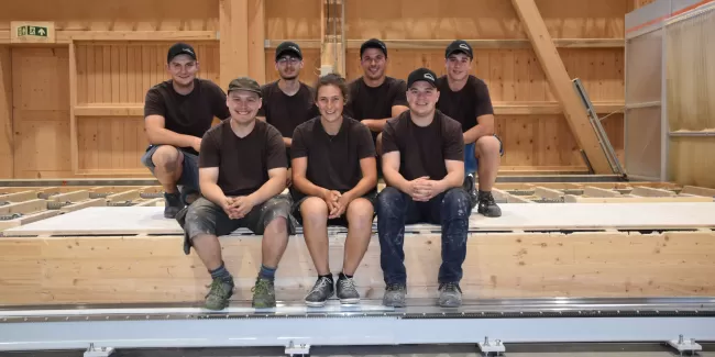 Sieben junge Zimmerleute in Arbeitskleidung posieren lächelnd in einer Werkhalle. Drei Personen sitzen auf einer Holzkonstruktion, vier knien oder stehen dahinter. Alle tragen dunkle T-Shirts und teils Kappen, im Hintergrund ist eine moderne Holzbauhalle sichtbar.