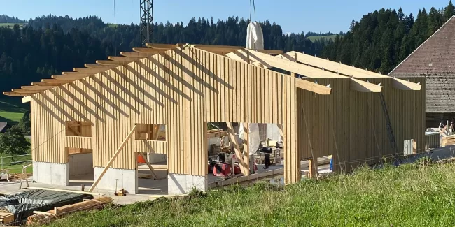 Ein modernes Holzbauprojekt von Zimmerei Hirschi AG: Im Vordergrund ist ein neu errichtetes Gebäude mit tragender Holzkonstruktion zu sehen. Ein Baukran hebt Materialien auf das Dach, während die klare Struktur der Holzbalken und Fassaden die Handwerkskunst hervorhebt. Umgeben von grünen Wiesen und bewaldeten Hügeln steht der Bau für nachhaltige, präzise und regionale Zimmermannsarbeit.