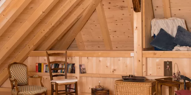 Gemütlicher Dachraum mit sichtbarer Holzbalkendecke, Holzboden und Einrichtung aus Stühlen, Bücherregal und warmem Licht.