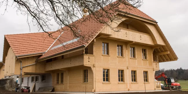 Ein neu erbautes Holzhaus mit roten Ziegeldächern und traditioneller Architektur steht auf einem ländlichen Grundstück; im Vordergrund ist Erde, im Hintergrund sind Wiesen und Bäume zu sehen.