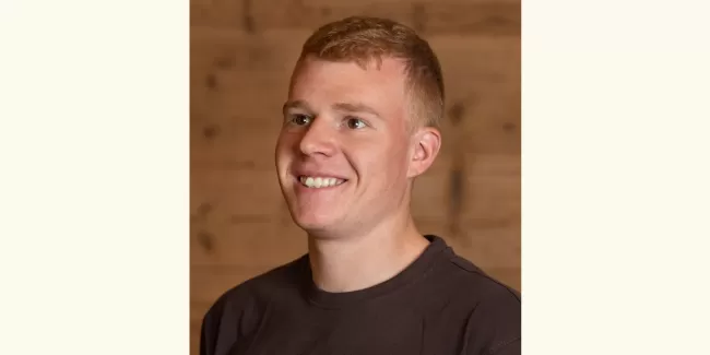 Porträt eines jungen Mannes mit kurzem, hellbraunem Haar, der lächelnd leicht nach rechts blickt. Er trägt ein dunkles T-Shirt, im Hintergrund ist eine warme Holzverkleidung zu sehen.