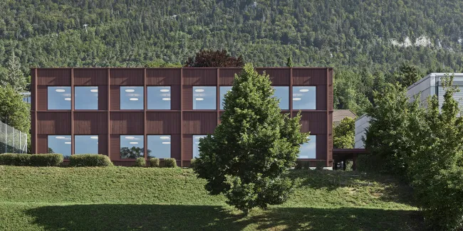 Aussenansicht eines modernen, zweigeschossigen Gebäudes mit dunkelbrauner Fassade und grossen, regelmässig angeordneten Fenstern. Das Gebäude steht in einer grünen Umgebung mit Wiese und Bäumen. Im Hintergrund erhebt sich ein bewaldeter Hügel unter klarem Himmel, was die ruhige, naturnahe Lage des Baus betont.