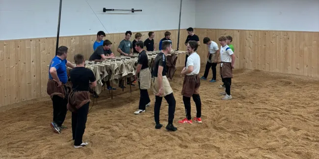 Innenaufnahme einer grossen Halle mit Holzdecke und Holzverkleidung an den Wänden. Mehrere junge Männer stehen in Arbeitskleidung auf einem mit Sägemehl bedeckten Boden und arbeiten gemeinsam an Holzbänken. Die Szene vermittelt eine handwerkliche Ausbildungs- oder Trainingssituation in einer Werk- oder Übungshalle.