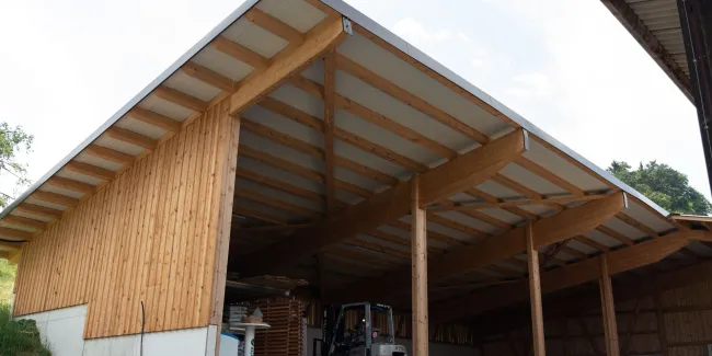 Aussenansicht einer grossen, offenen Lager- oder Maschinenhalle in Holzbauweise mit geneigtem Dach. Die Konstruktion aus sichtbaren Holzbalken ruht auf einem Betonsockel. Im Inneren der Halle sind ein Gabelstapler und gelagerte Materialien zu erkennen. Die Umgebung wirkt ländlich und funktional gestaltet.