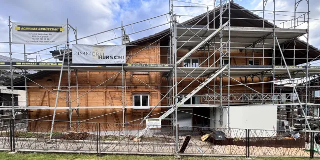 Holzhaus im Bau mit eingerüsteter Fassade und Baustellenbanner der Zimmerei Hirschi unter blauem Himmel.