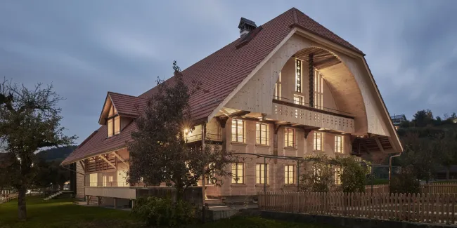 Traditionelles Holzhaus im Emmental in der Abenddämmerung, mit geschwungenem Giebeldach und aufwendig gestalteter Holzfassade. Warmes Licht fällt aus den Fenstern und betont die handwerklichen Details der historischen Holzkonstruktion. Das Gebäude steht in einer ruhigen, ländlichen Umgebung mit Garten, Bäumen und Holzzaun.