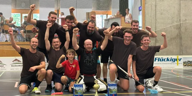 Eine Unihockey-Mannschaft feiert gemeinsam mit einem Kind und einem Torhüter auf dem Hallenboden einen Turniersieg. Vor der Gruppe stehen ein Pokal und zwei Getränkepreise.