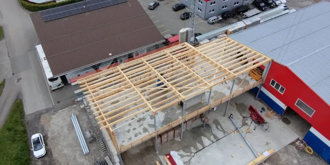 Luftaufnahme eines Gewerbebaus während der Bauphase. Zu sehen ist die im Aufbau befindliche Holzdachkonstruktion mit Tragwerk und Sparren auf einem Industriegebäude, umgeben von Werkhallen und Baustelleneinrichtungen.