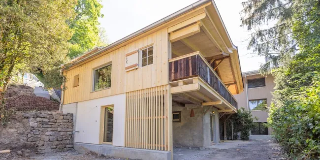 einblicke in ein holzbauprojekt in gunten: die aussenansicht zeigt ein haus mit heller holzfassade und balkon, eingebettet zwischen bäumen und einem steinmäuerchen. im inneren prägen helle holzdecken und sichtbare balken, grosse fenster und warme holzoberflächen die räume. zu sehen sind ein grosszügiger wohnbereich mit zugang zum balkon und ausblick ins grüne, eine holzküche mit arbeitsinsel sowie ein badezimmer mit rundem spiegel und aufgesetztem waschbecken. helles ziegelmauerwerk und sorgfältig erhaltene