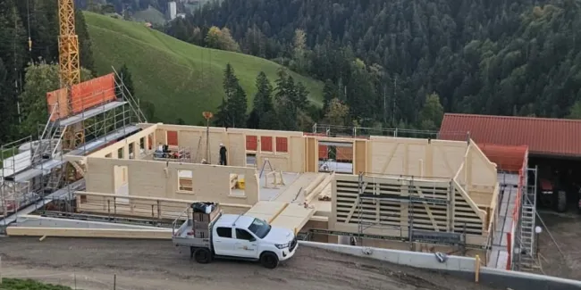 Holzbau im Rohbau mit Baukran an einem grünen Hang, mit bewaldeten Hügeln und schneebedeckten Bergen im Hintergrund.