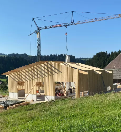 Ein modernes Holzbauprojekt von Zimmerei Hirschi AG: Im Vordergrund ist ein neu errichtetes Gebäude mit tragender Holzkonstruktion zu sehen. Ein Baukran hebt Materialien auf das Dach, während die klare Struktur der Holzbalken und Fassaden die Handwerkskunst hervorhebt. Umgeben von grünen Wiesen und bewaldeten Hügeln steht der Bau für nachhaltige, präzise und regionale Zimmermannsarbeit.