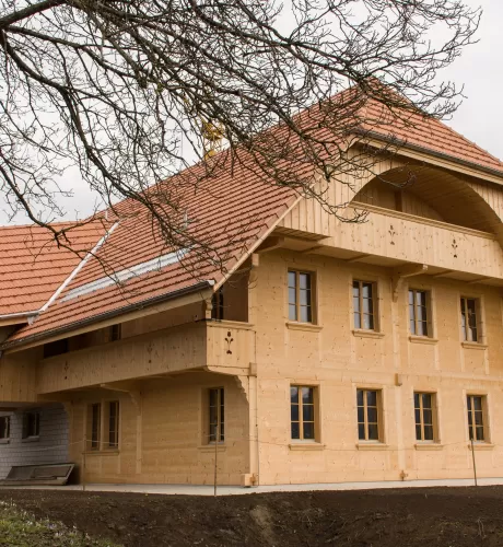 Ein neu erbautes Holzhaus mit roten Ziegeldächern und traditioneller Architektur steht auf einem ländlichen Grundstück; im Vordergrund ist Erde, im Hintergrund sind Wiesen und Bäume zu sehen.