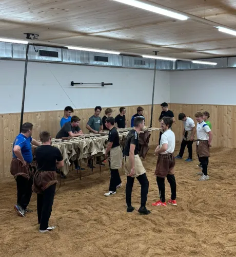 Innenaufnahme einer grossen Halle mit Holzdecke und Holzverkleidung an den Wänden. Mehrere junge Männer stehen in Arbeitskleidung auf einem mit Sägemehl bedeckten Boden und arbeiten gemeinsam an Holzbänken. Die Szene vermittelt eine handwerkliche Ausbildungs- oder Trainingssituation in einer Werk- oder Übungshalle.