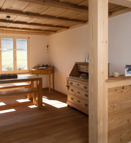 Innenansicht eines wohnlichen Ess- und Arbeitsbereichs in Holzbauweise. Sichtbare Holzbalken an der Decke, ein massiver Holztisch mit Bänken sowie ein Holzschreibtisch stehen in einem hellen Raum mit Holzboden. Tageslicht fällt durch ein Fenster ein und betont die warme, natürliche Atmosphäre sowie die handwerklich ausgeführten Holzdetails.