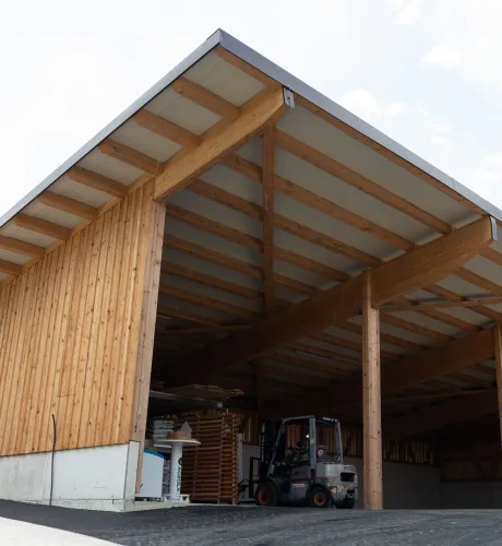 Aussenansicht einer grossen, offenen Lager- oder Maschinenhalle in Holzbauweise mit geneigtem Dach. Die Konstruktion aus sichtbaren Holzbalken ruht auf einem Betonsockel. Im Inneren der Halle sind ein Gabelstapler und gelagerte Materialien zu erkennen. Die Umgebung wirkt ländlich und funktional gestaltet.