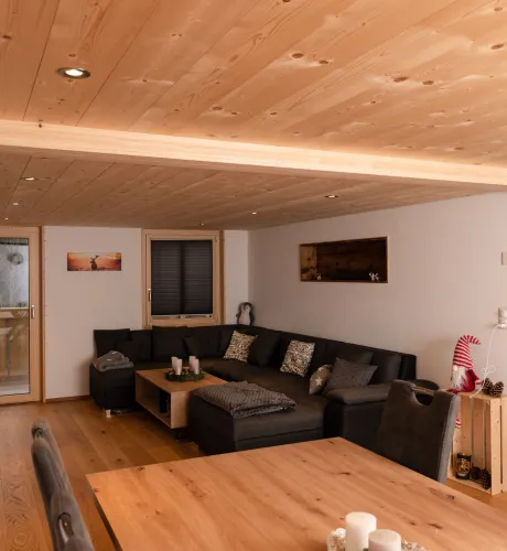 Innenansicht eines gemütlichen Wohnzimmers mit hellem Holzboden und sichtbarer Holzdecke. An der Wand hängt ein Fernseher über einem niedrigen Sideboard. Mehrere Holzfenster geben den Blick in eine bewaldete Landschaft frei. Rechts führt eine Glastür in einen angrenzenden Raum, der Wohnbereich ist warm und wohnlich gestaltet.