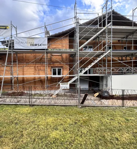 Holzhaus im Bau mit eingerüsteter Fassade und Baustellenbanner der Zimmerei Hirschi unter blauem Himmel.