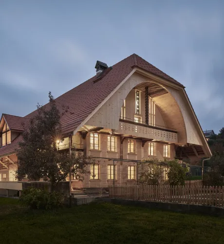 Traditionelles Holzhaus im Emmental in der Abenddämmerung, mit geschwungenem Giebeldach und aufwendig gestalteter Holzfassade. Warmes Licht fällt aus den Fenstern und betont die handwerklichen Details der historischen Holzkonstruktion. Das Gebäude steht in einer ruhigen, ländlichen Umgebung mit Garten, Bäumen und Holzzaun.