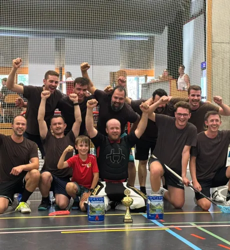 Eine Unihockey-Mannschaft feiert gemeinsam mit einem Kind und einem Torhüter auf dem Hallenboden einen Turniersieg. Vor der Gruppe stehen ein Pokal und zwei Getränkepreise.