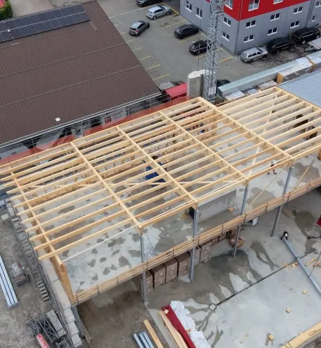 Luftaufnahme eines Gewerbebaus während der Bauphase. Zu sehen ist die im Aufbau befindliche Holzdachkonstruktion mit Tragwerk und Sparren auf einem Industriegebäude, umgeben von Werkhallen und Baustelleneinrichtungen.