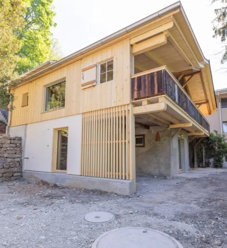 einblicke in ein holzbauprojekt in gunten: die aussenansicht zeigt ein haus mit heller holzfassade und balkon, eingebettet zwischen bäumen und einem steinmäuerchen. im inneren prägen helle holzdecken und sichtbare balken, grosse fenster und warme holzoberflächen die räume. zu sehen sind ein grosszügiger wohnbereich mit zugang zum balkon und ausblick ins grüne, eine holzküche mit arbeitsinsel sowie ein badezimmer mit rundem spiegel und aufgesetztem waschbecken. helles ziegelmauerwerk und sorgfältig erhaltene