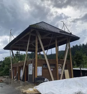 Modernes Holzgebäude mit markantem, erhöhtem Satteldach und offener Konstruktion, aufgenommen an einem bewölkten Tag inmitten einer grünen Hügellandschaft.
