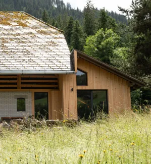 Aussenansicht eines umgebauten Bauernhauses in Holzbauweise mit traditionellem Satteldach, teilweise mit Moos bewachsen. Der Anbau aus hellem Holz fügt sich an den bestehenden Bau an. Im Vordergrund eine Wiese mit hohem Gras, im Hintergrund ein bewaldeter Hang.