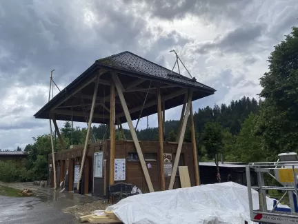 Modernes Holzgebäude mit markantem, erhöhtem Satteldach und offener Konstruktion, aufgenommen an einem bewölkten Tag inmitten einer grünen Hügellandschaft.
