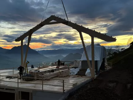Holzbauarbeiten auf einer Bergbaustelle bei Sonnenaufgang – Zimmerleute montieren eine Dachkonstruktion mit Blick auf die umliegenden Berge und das Tal unter einem farbenprächtigen Morgenhimmel.