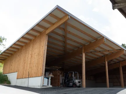 Aussenansicht einer grossen, offenen Lager- oder Maschinenhalle in Holzbauweise mit geneigtem Dach. Die Konstruktion aus sichtbaren Holzbalken ruht auf einem Betonsockel. Im Inneren der Halle sind ein Gabelstapler und gelagerte Materialien zu erkennen. Die Umgebung wirkt ländlich und funktional gestaltet.