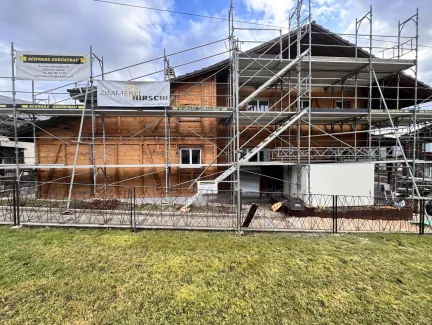 Holzhaus im Bau mit eingerüsteter Fassade und Baustellenbanner der Zimmerei Hirschi unter blauem Himmel.