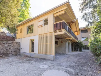 einblicke in ein holzbauprojekt in gunten: die aussenansicht zeigt ein haus mit heller holzfassade und balkon, eingebettet zwischen bäumen und einem steinmäuerchen. im inneren prägen helle holzdecken und sichtbare balken, grosse fenster und warme holzoberflächen die räume. zu sehen sind ein grosszügiger wohnbereich mit zugang zum balkon und ausblick ins grüne, eine holzküche mit arbeitsinsel sowie ein badezimmer mit rundem spiegel und aufgesetztem waschbecken. helles ziegelmauerwerk und sorgfältig erhaltene