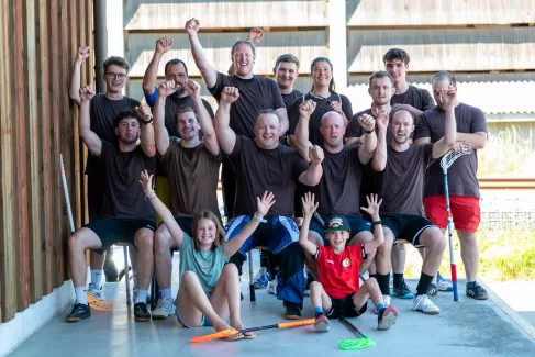 Eine fröhliche Gruppe von 17 Personen – Männer, eine Frau und zwei Kinder – posiert jubelnd für ein Gruppenfoto. Alle tragen Sportkleidung, viele einheitlich braune T-Shirts, und einige halten Unihockeyschläger in der Hand. Die Stimmung ist ausgelassen, viele recken die Arme in die Höhe. Im Vordergrund sitzen zwei Kinder mit hochgestreckten Armen, im Hintergrund ist ein Holzgebäude und Sonnenlicht zu sehen – ein sportlicher Team-Moment voller Energie.