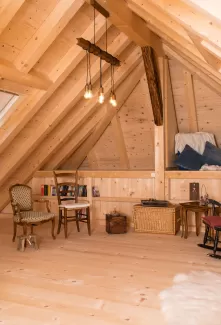 Gemütlicher Dachraum mit sichtbarer Holzbalkendecke, Holzboden und Einrichtung aus Stühlen, Bücherregal und warmem Licht.